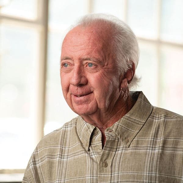 An older man in a plaid shirt standing in front of a window.